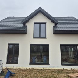 PRYM - Dwukondygnacyjny dom z jasną elewacją, grafitowym dachem i oknami, widoczne ślady prac wykończeniowych na zewnątrz, w oknach odbijają się worki z materiałami budowlanymi i sylwetka człowieka.