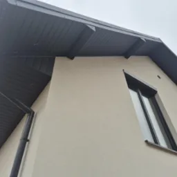 Fragment elewacji domu z nowym ociepleniem i grafitowym orynnowaniem, widok z dołu na narożnik budynku i okna.