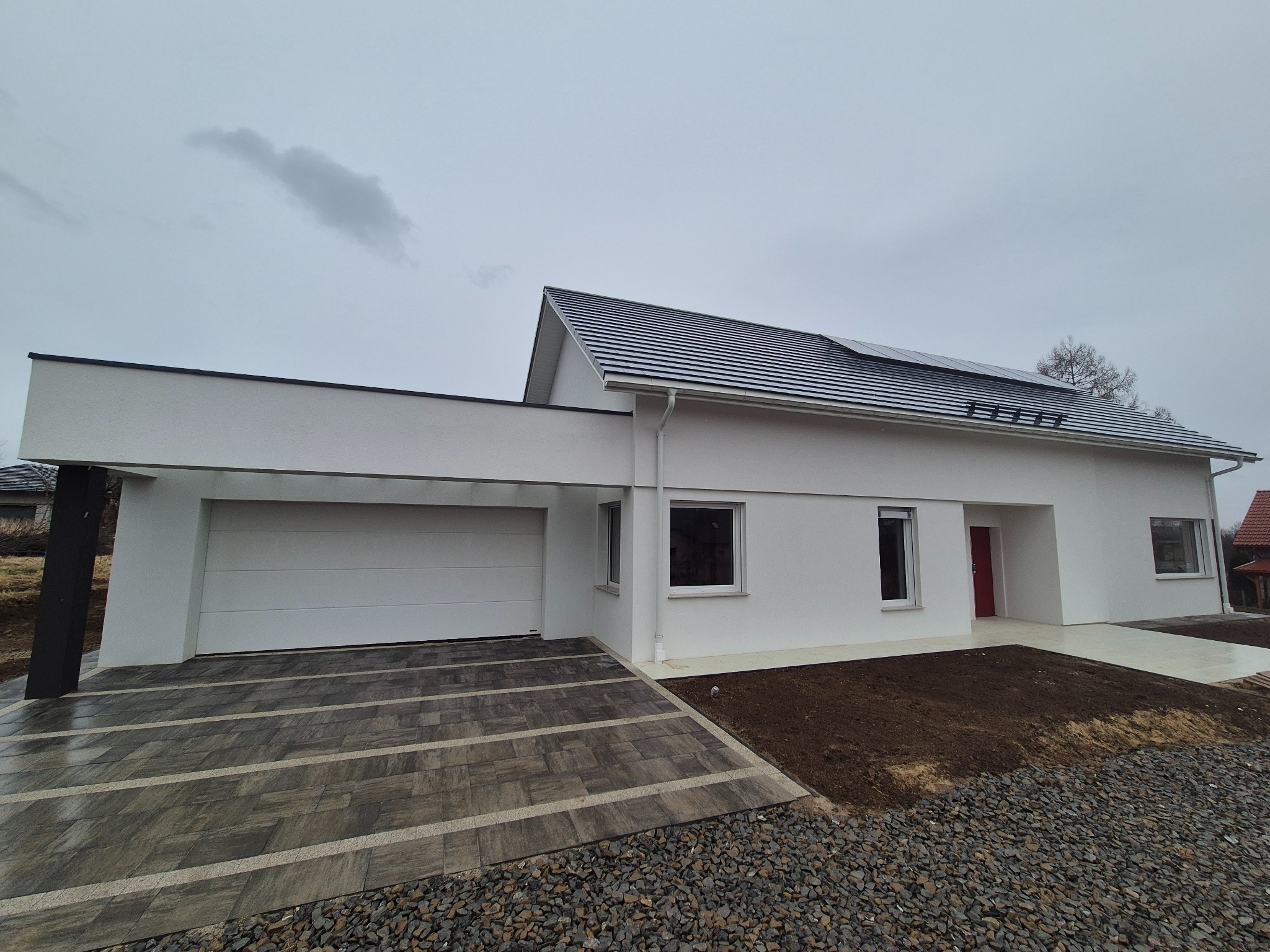 Nowoczesny, parterowy dom z białymi ścianami, ciemną dachówką i czerwoną stolarką drzwiową, widok z zewnątrz na elewację i podjazd wyłożony kostką brukową w odcieniach szarości, pochmurne niebo w tle.