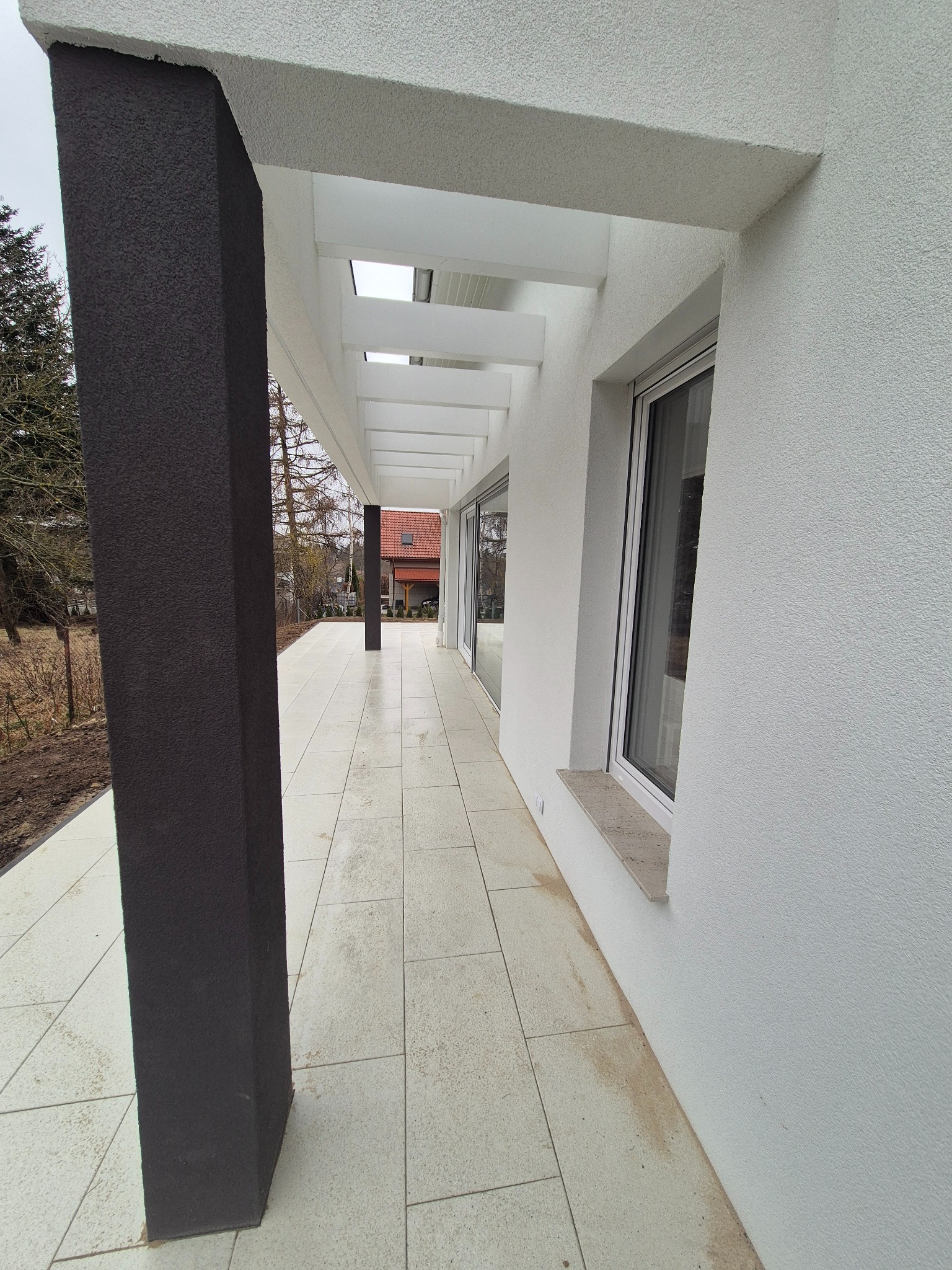 Nowoczesny taras z białymi ścianami o fakturze baranka, ciemnymi filarami i jasną, kwadratową nawierzchnią z płytek. Widoczne okno z jasnym parapetem i zadaszenie z białych belek.