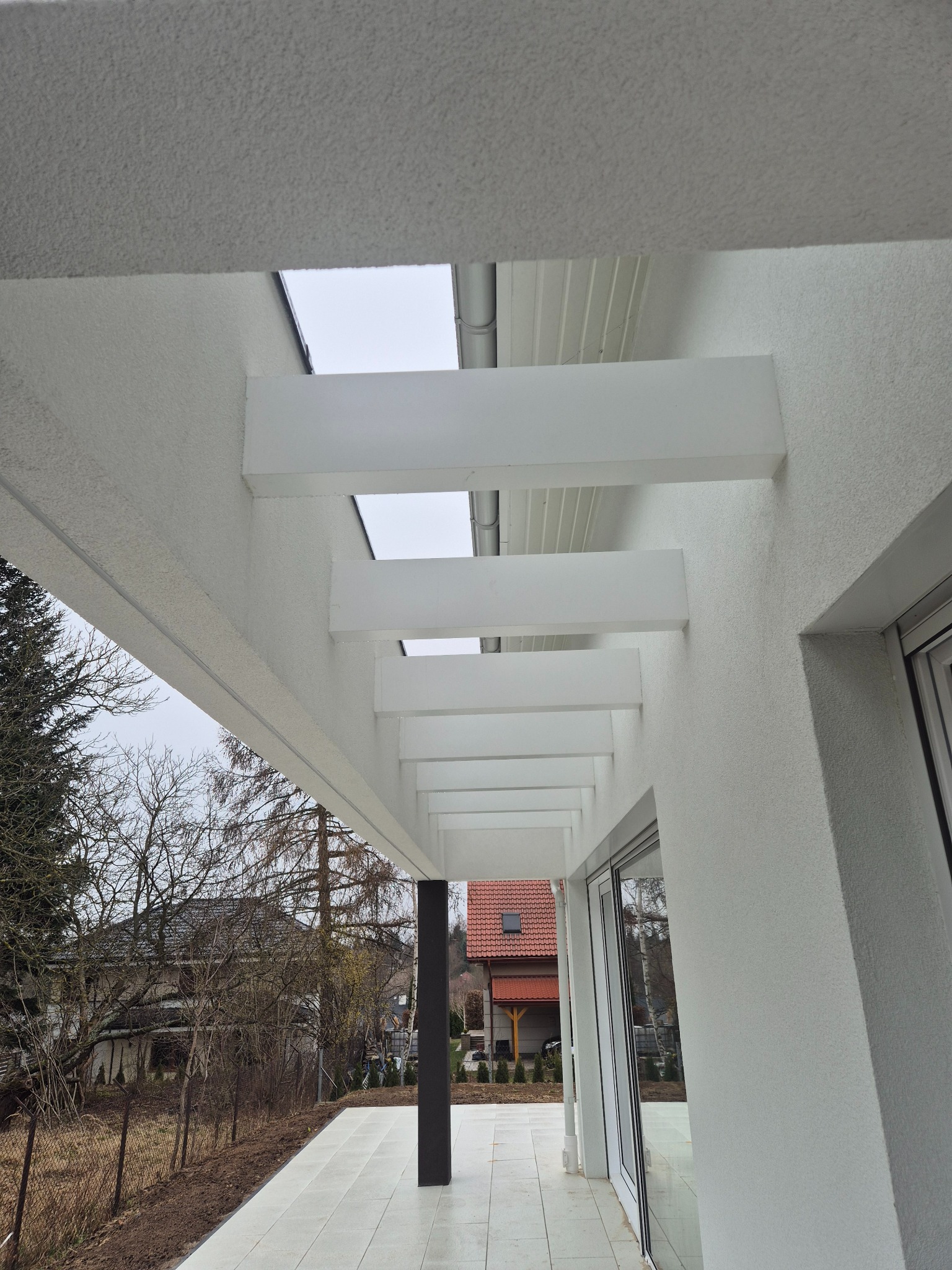 Nowoczesny taras z białymi, minimalistycznymi elementami konstrukcyjnymi i widokiem na ogród w pochmurny dzień.