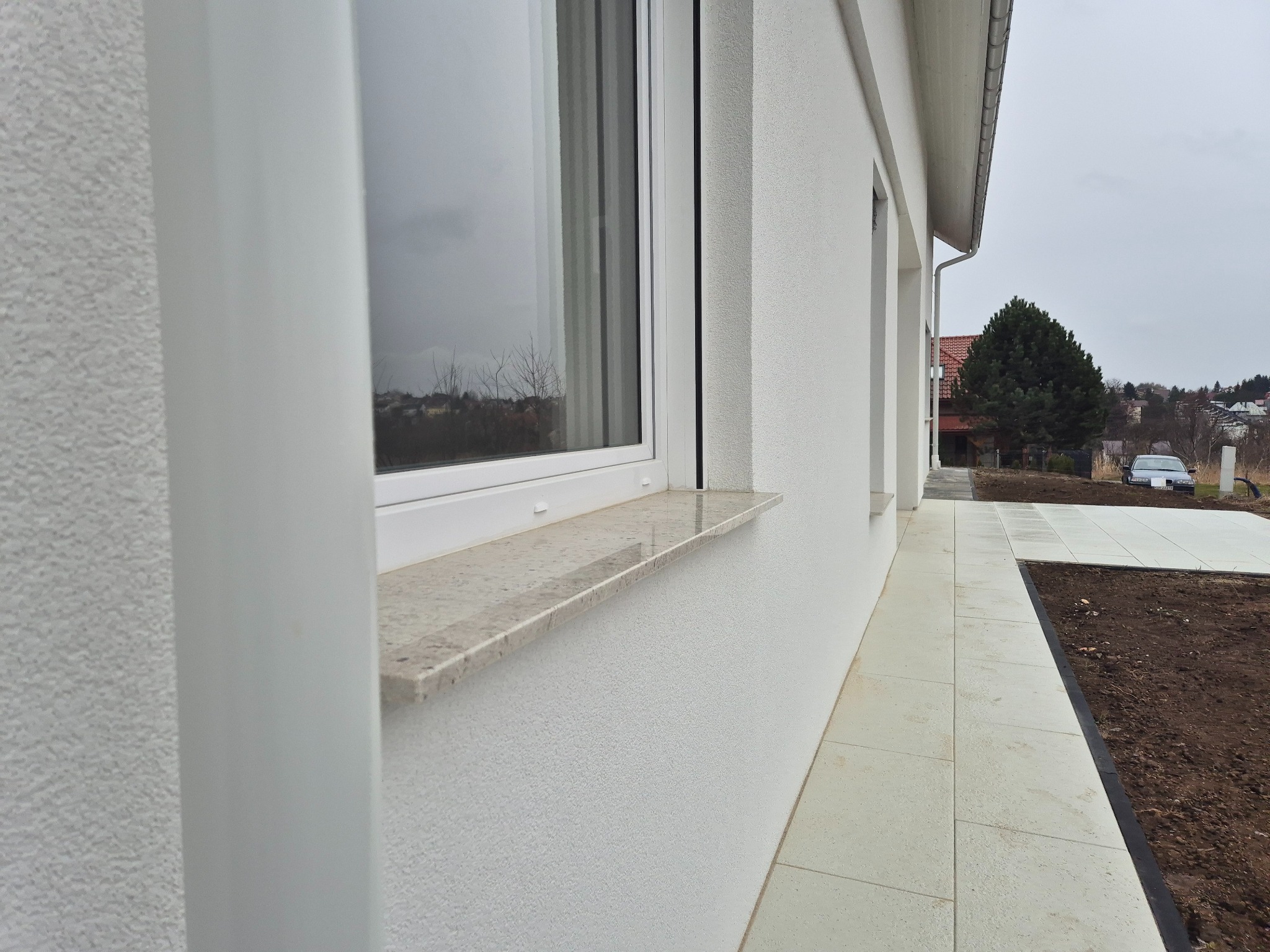Biała elewacja domu z widocznym oknem z marmurowym parapetem i jasną kostką brukową na zewnątrz, w tle zabudowa jednorodzinna i pochmurne niebo.