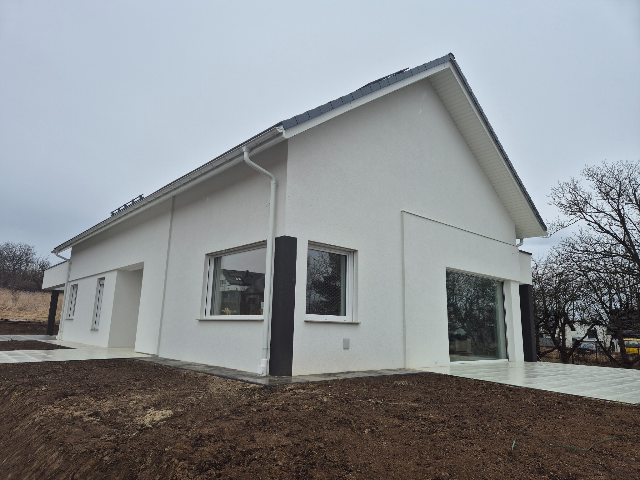 Nowoczesny, parterowy dom z białą elewacją i dużymi oknami, czarnymi akcentami kolumn i ram okiennych, szarą dachówką, widok z zewnątrz na świeżo przygotowany teren pod ogród