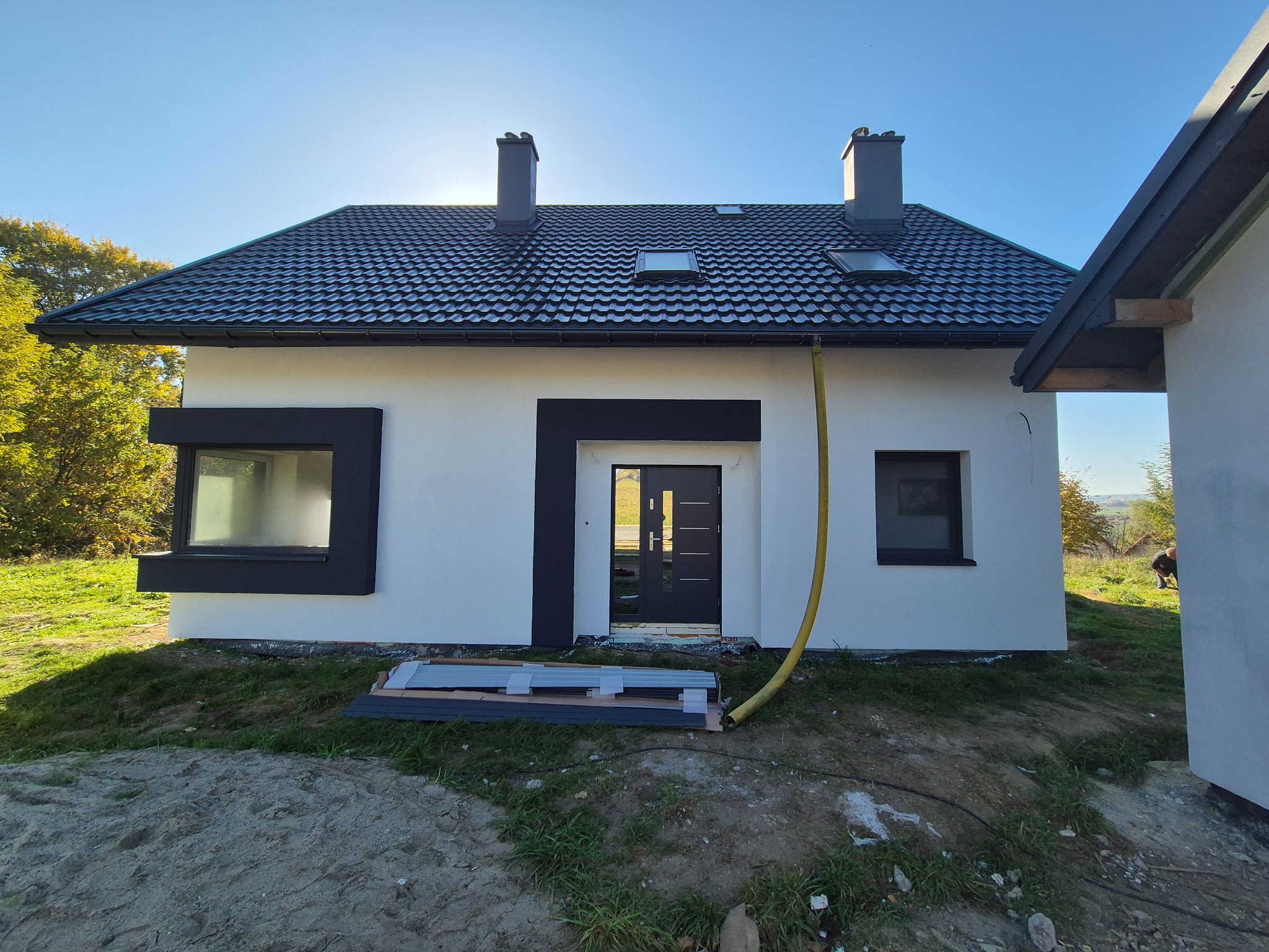 Nowoczesny dom jednorodzinny z białą elewacją i czarnymi obramowaniami okien i drzwi, widoczne rury i materiały budowlane na trawniku przed domem, dach pokryty czarną dachówką.