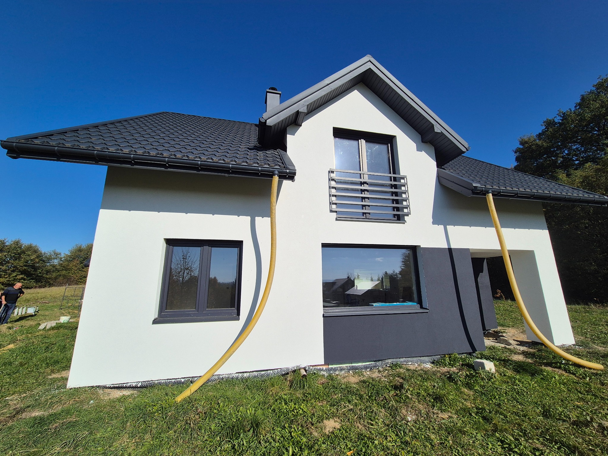 Nowoczesny dom z białą elewacją, czarną stolarką okienną i dachem z blachodachówki, z widocznymi żółtymi wężami do ocieplania, stojący na trawiastym terenie.