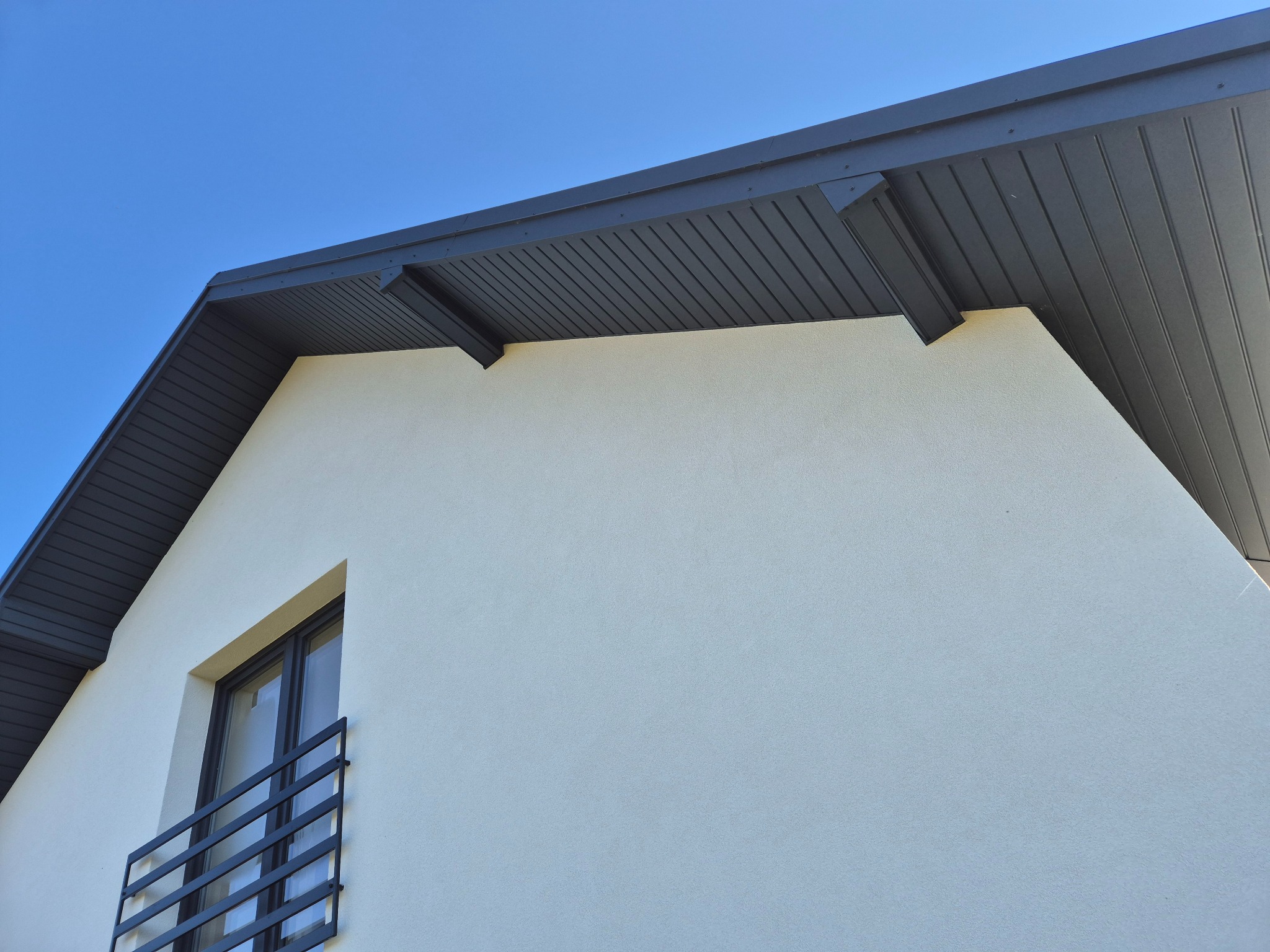 Fragment ocieplonej elewacji budynku z szarą podbitką dachową i balkonem z czarną balustradą na tle błękitnego nieba.