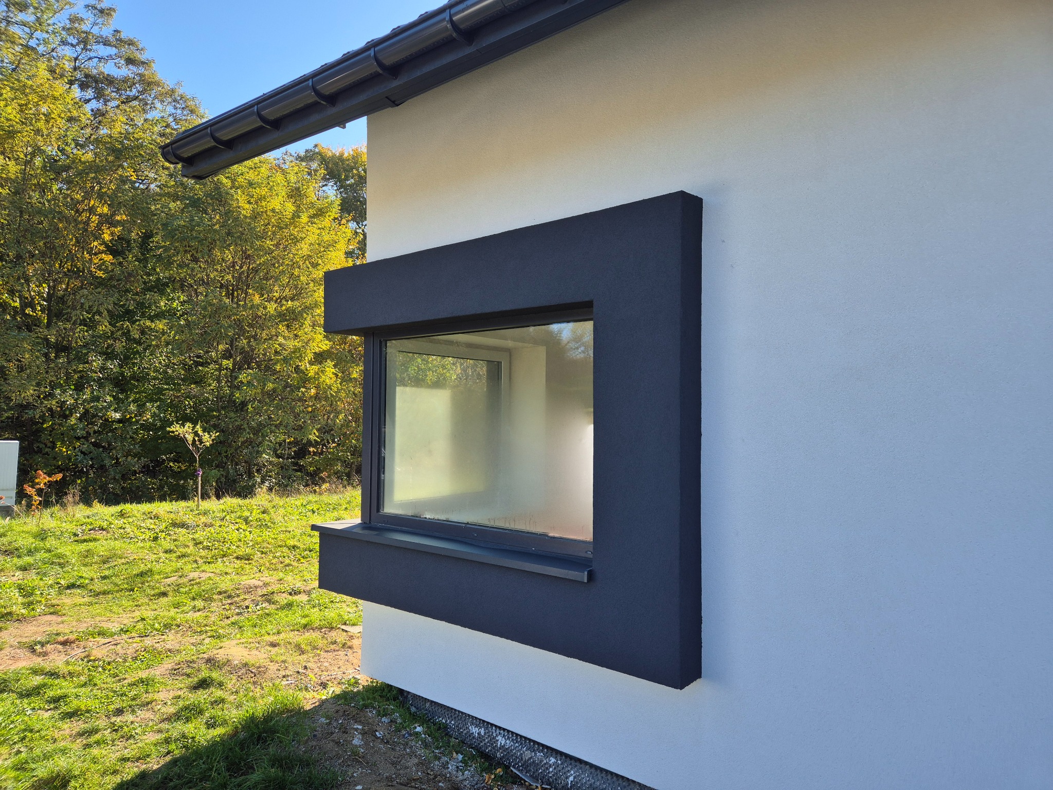 Nowoczesne okno z ciemną, grubą ramą wystającą poza elewację budynku o jasnej, gładkiej strukturze. W tle widoczne drzewa i trawa.