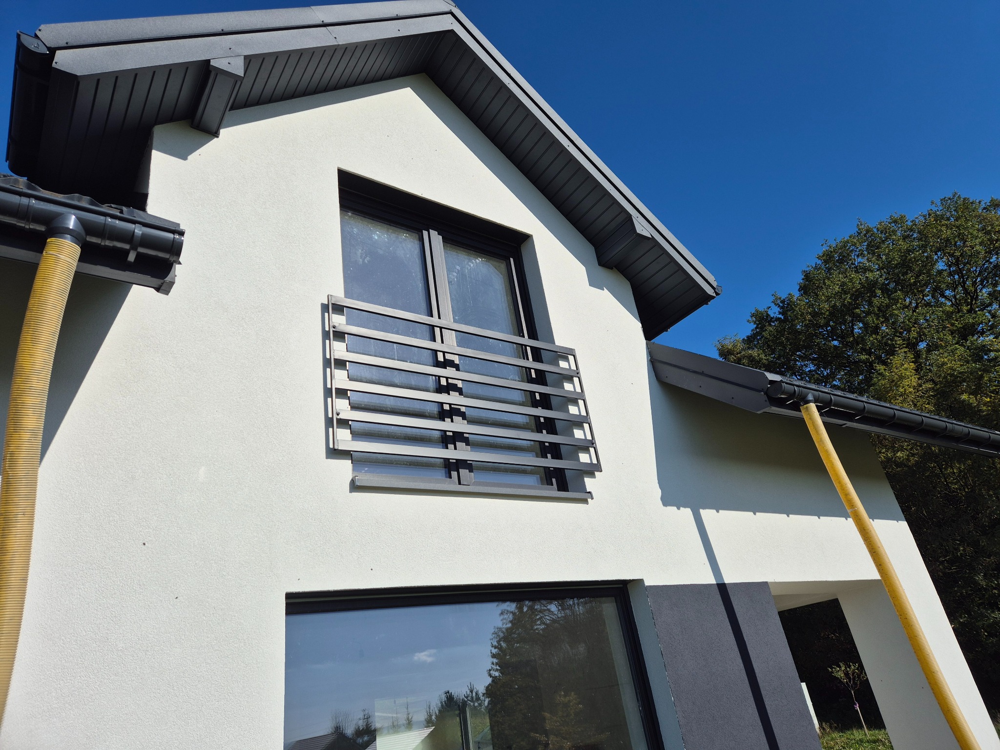 Nowoczesny dom z białą elewacją i szarymi detalami, widoczna żółta rura do podawania materiału elewacyjnego.