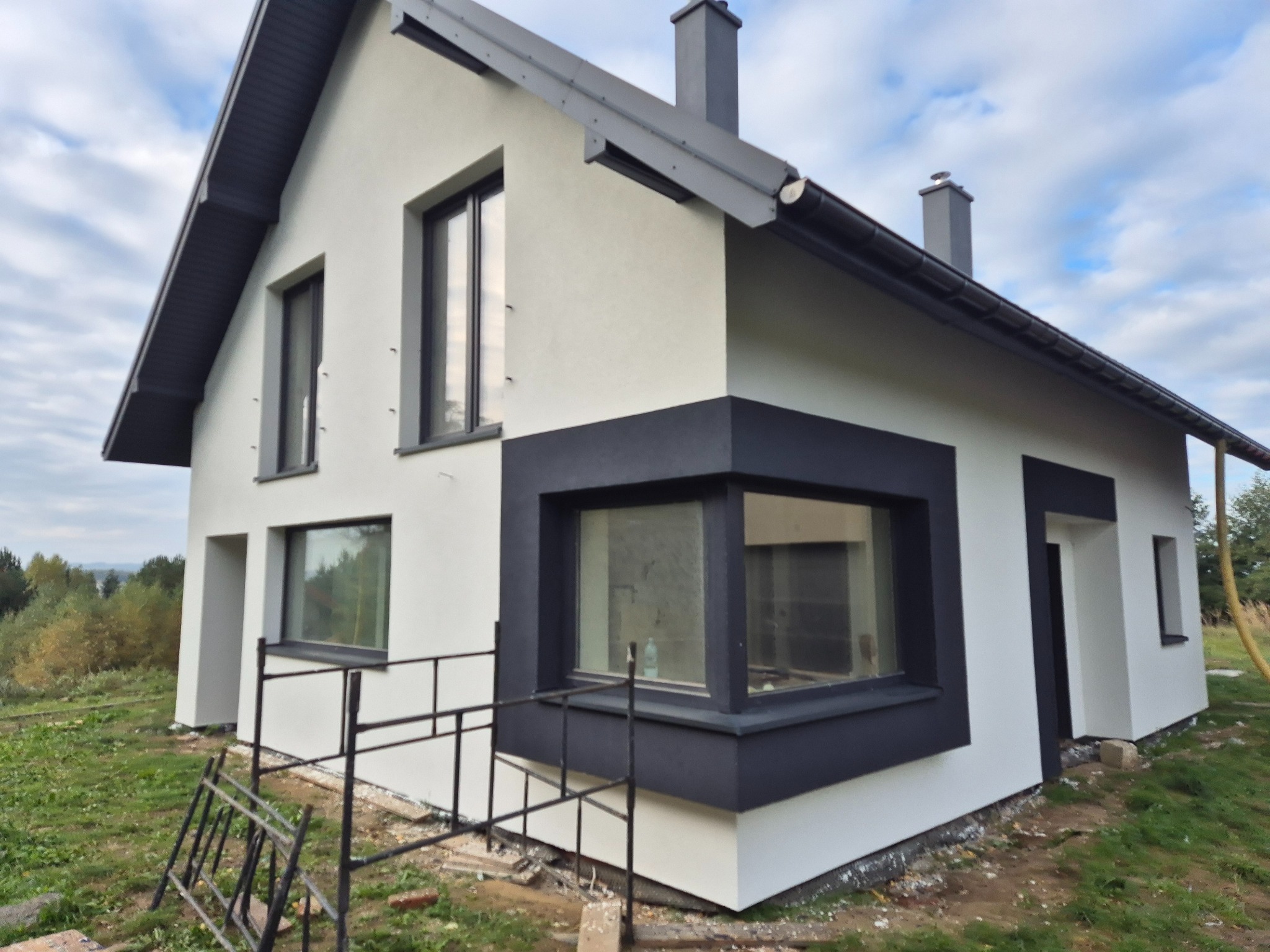 Nowoczesny dom jednorodzinny z białą elewacją i grafitowymi detalami okiennymi oraz dachem, widoczny z boku, na trawniku leżą resztki materiałów budowlanych oraz drabina.