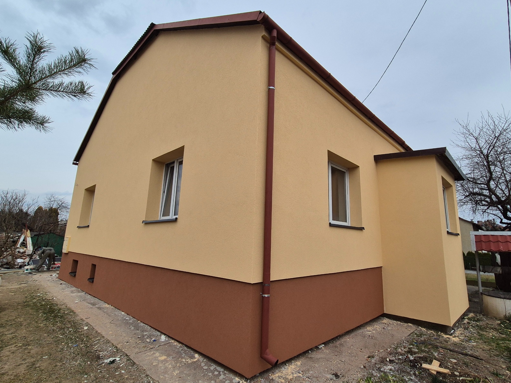 Dwukolorowa elewacja domu jednorodzinnego z brązowym cokołem, beżową górną częścią, brązową rynną i białymi ramami okiennymi, widziana pod kątem w pochmurny dzień.