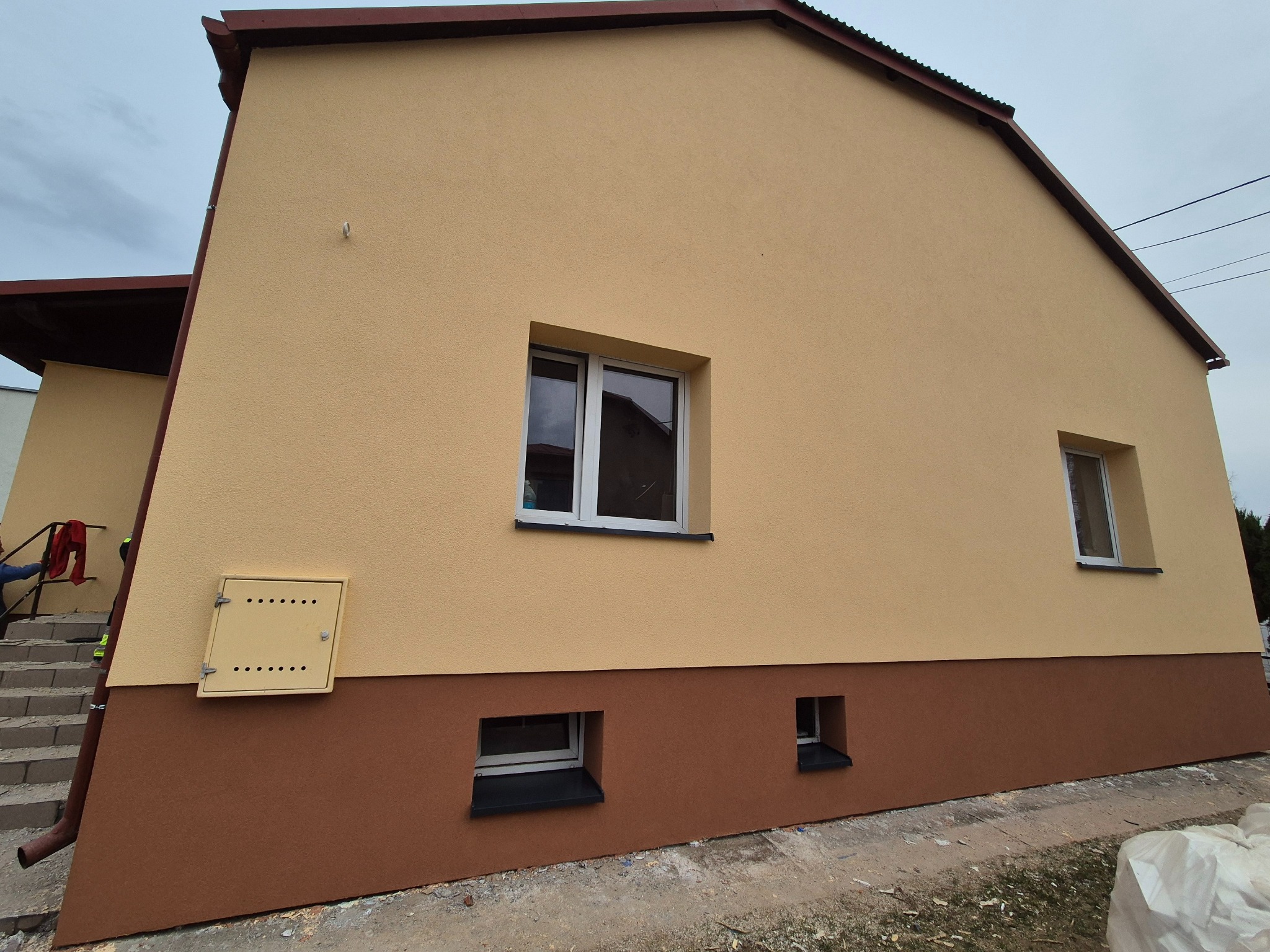 Ocieplona elewacja domu jednorodzinnego z brązowym cokołem, widoczne okna piwniczne z czarnymi parapetami, skrzynka gazowa z otworami wentylacyjnymi w kolorze elewacji.