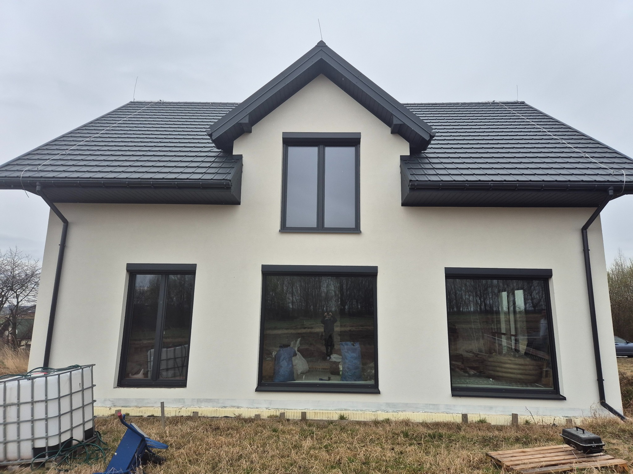 Dwukondygnacyjny dom z jasną elewacją, grafitowym dachem i oknami, widoczne ślady prac wykończeniowych na zewnątrz, w oknach odbijają się worki z materiałami budowlanymi i sylwetka człowieka.