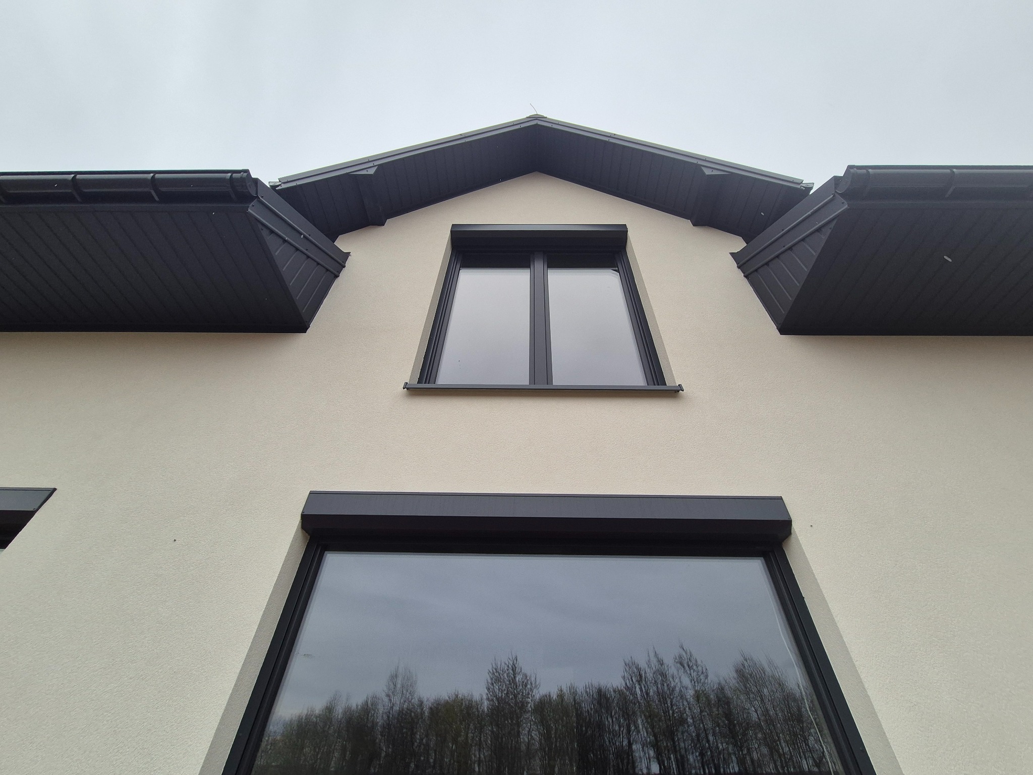 Elewacja domu z jasnym tynkiem i ciemnymi elementami wykończeniowymi, widok z dołu, okna z roletami zewnętrznymi.