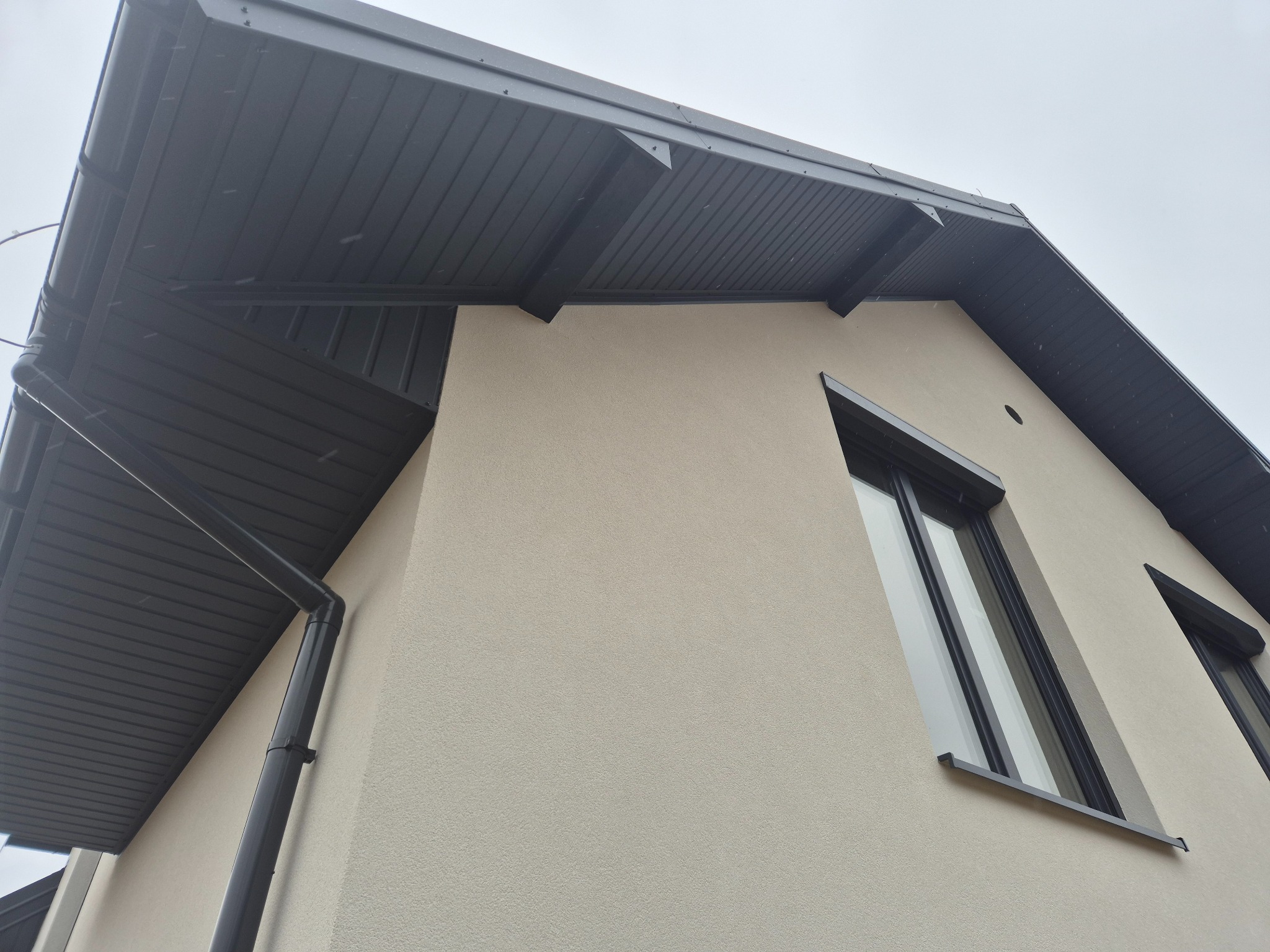 Fragment elewacji domu z nowym ociepleniem i grafitowym orynnowaniem, widok z dołu na narożnik budynku i okna.