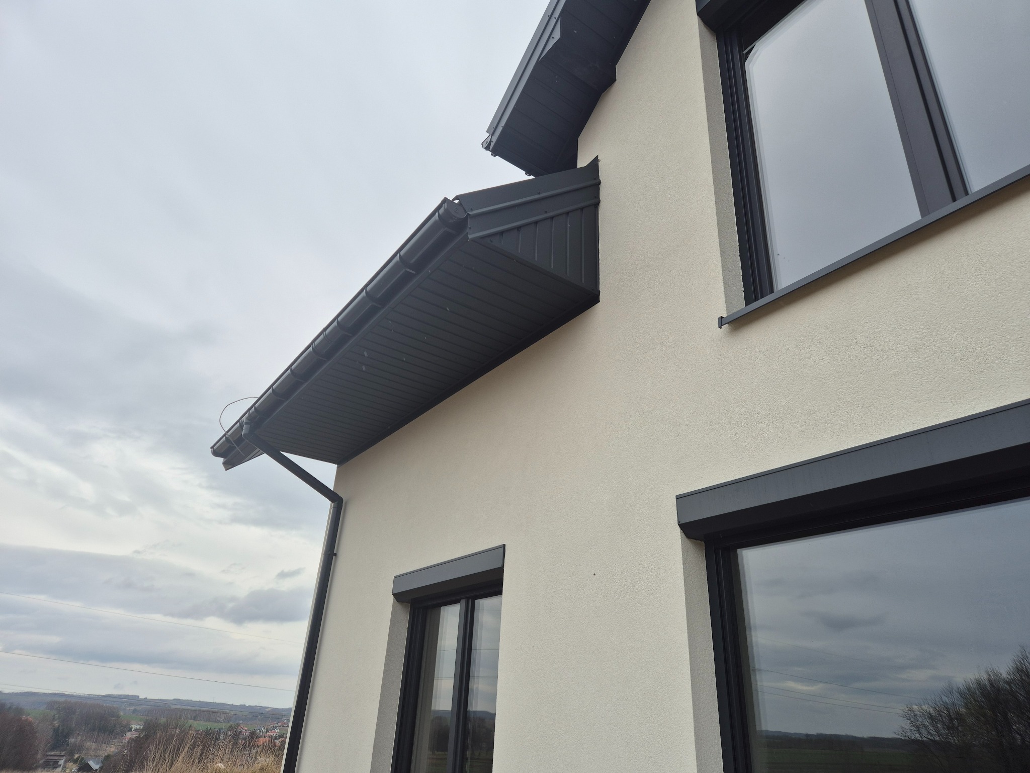 Fragment elewacji domu z oknami z szarymi roletami zewnętrznymi, widoczny system rynnowy i struktura tynku baranka.