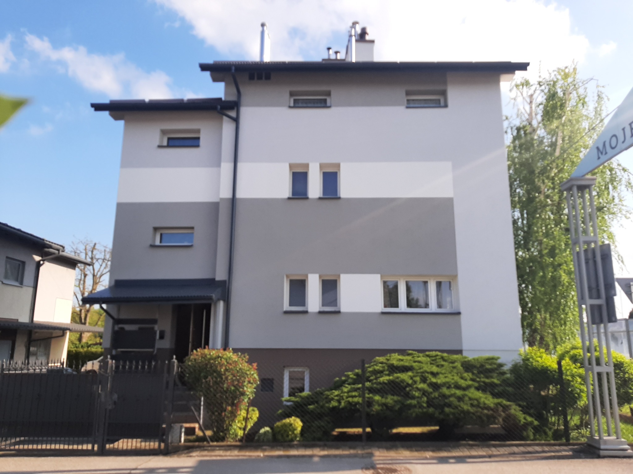 Dwupiętrowy dom z elewacją w odcieniach szarości i bieli, widoczny z poziomu ulicy, z fragmentem ogrodzenia i zielenią w tle, oświetlony porannym słońcem.