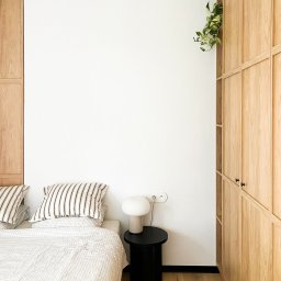 othe.studio - Minimalistyczna sypialnia z zabudową szafy z jasnego drewna, lampką na czarnym stoliku nocnym i rośliną pnącą się z półki.