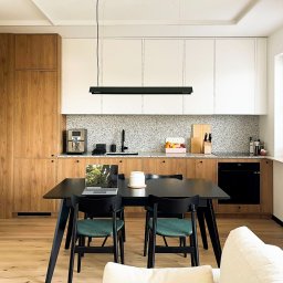 othe.studio - Nowoczesna kuchnia z drewnianymi i białymi szafkami, szarym blatem, czarnym stołem jadalnym i krzesłami z zielonymi siedziskami, laptop na stole, widok z salonu.