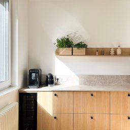 othe.studio - Jasna kuchnia z drewnianymi szafkami, czarnymi okrągłymi uchwytami, ekspres do kawy Smeg, czajnik Smeg, półka z ziołami i dekoracjami, parkiet w jodełkę.