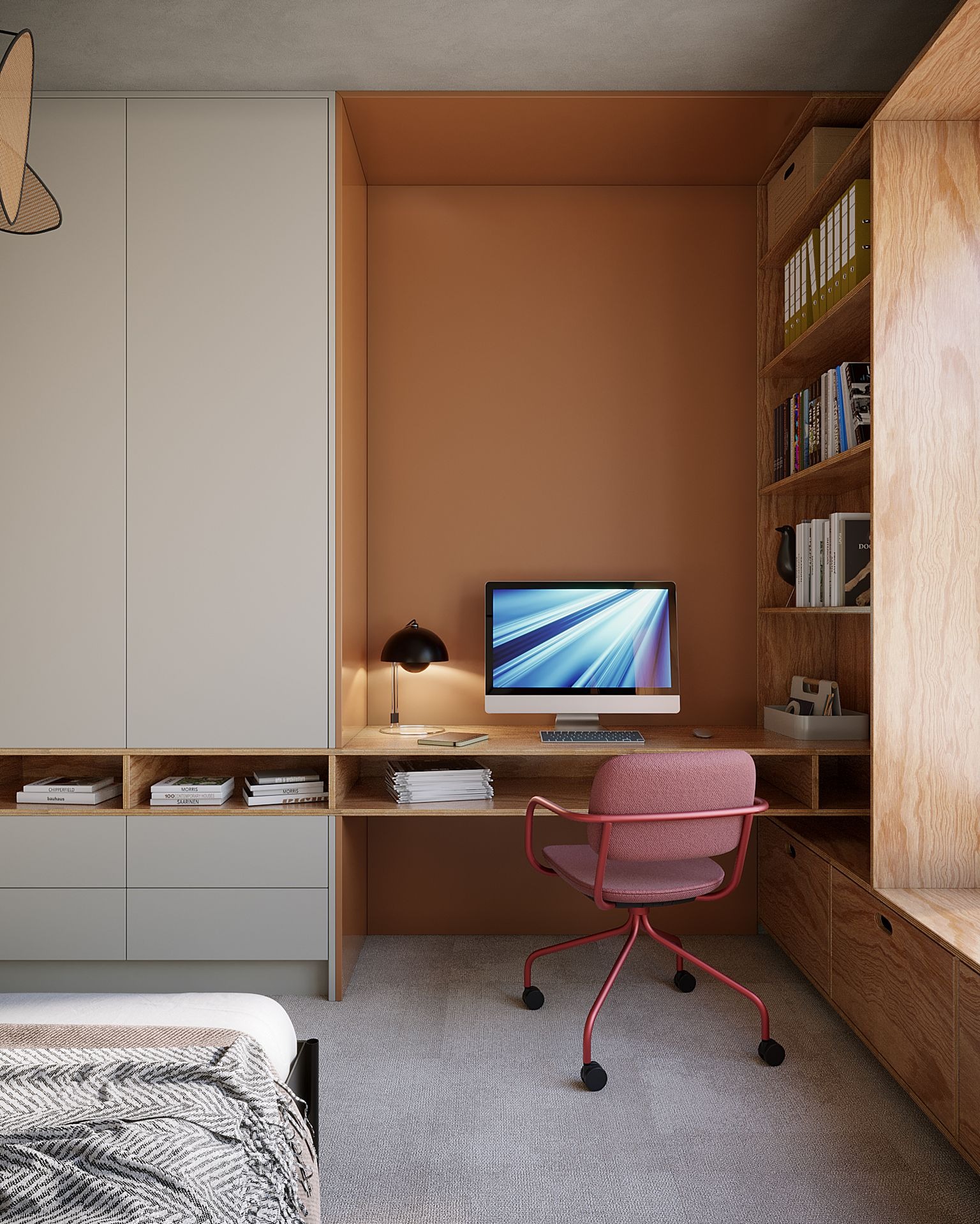 Pokój z biurkiem wbudowanym w szafę, komputerem, krzesłem i regałem z książkami, oświetlony lampką biurkową, w tle łóżko z narzutą.