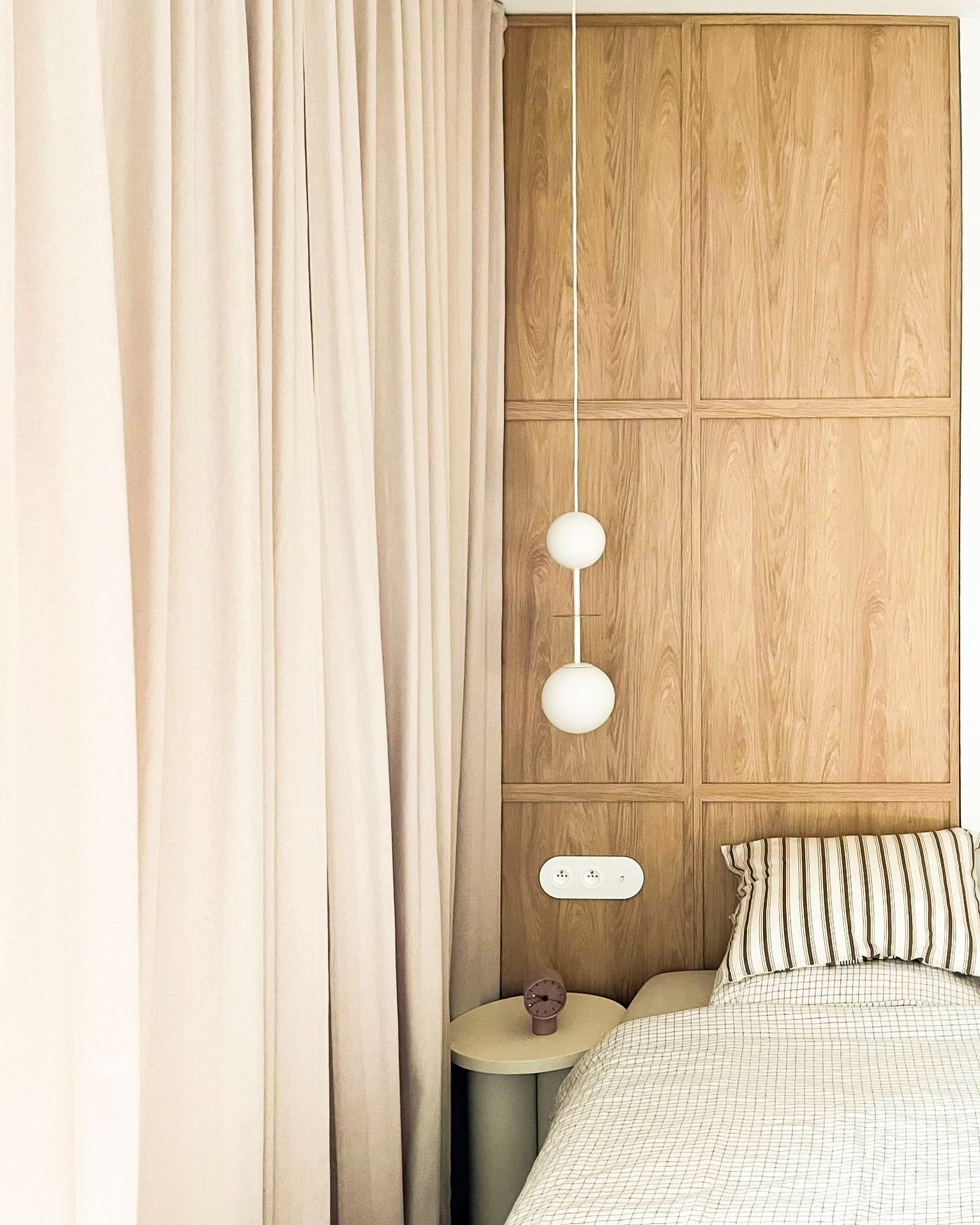 Sypialnia z drewnianą zabudową ścienną, lampą kulistą, beżowymi zasłonami i stolikiem nocnym z fioletowym zegarem.