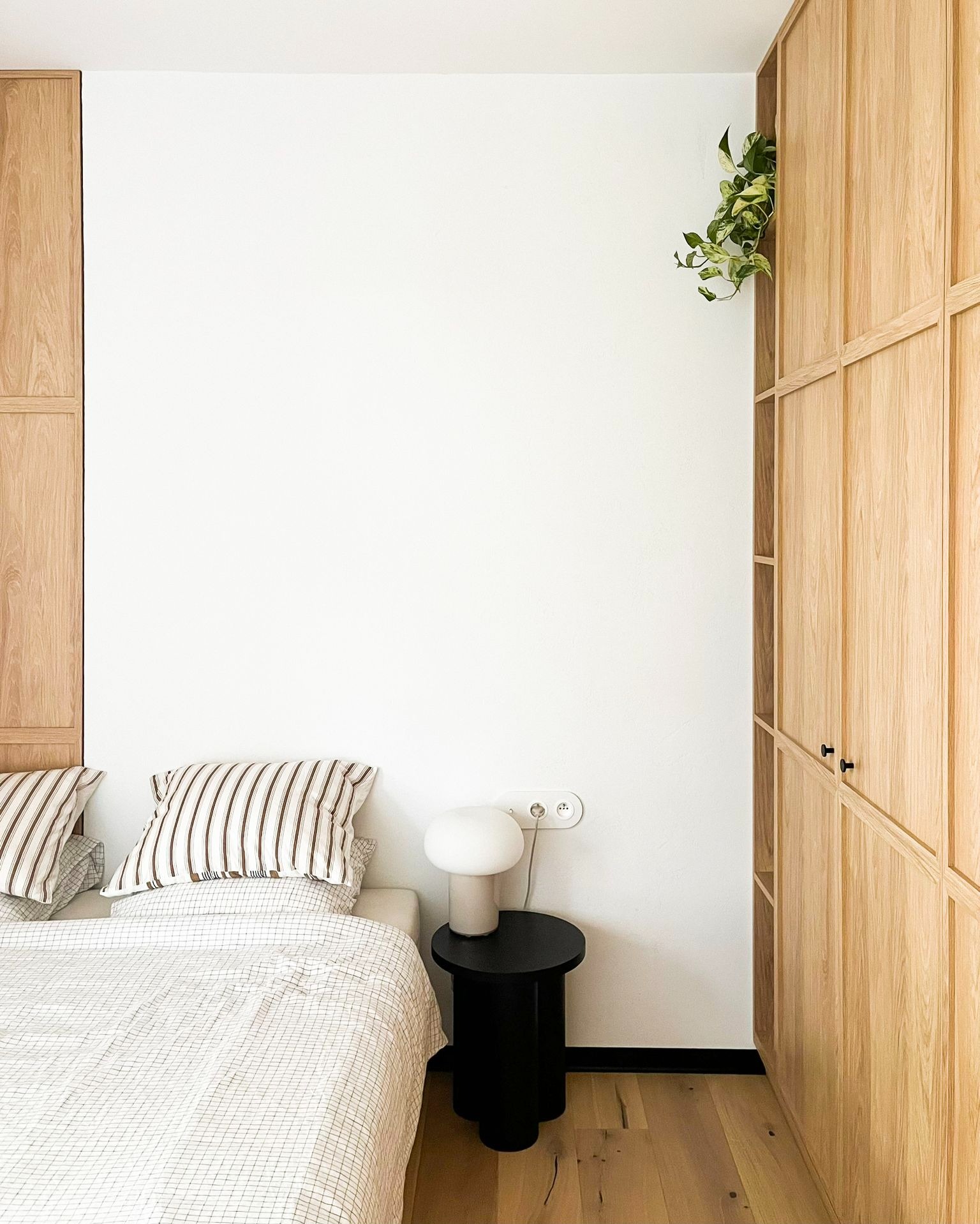 Minimalistyczna sypialnia z zabudową szafy z jasnego drewna, lampką na czarnym stoliku nocnym i rośliną pnącą się z półki.