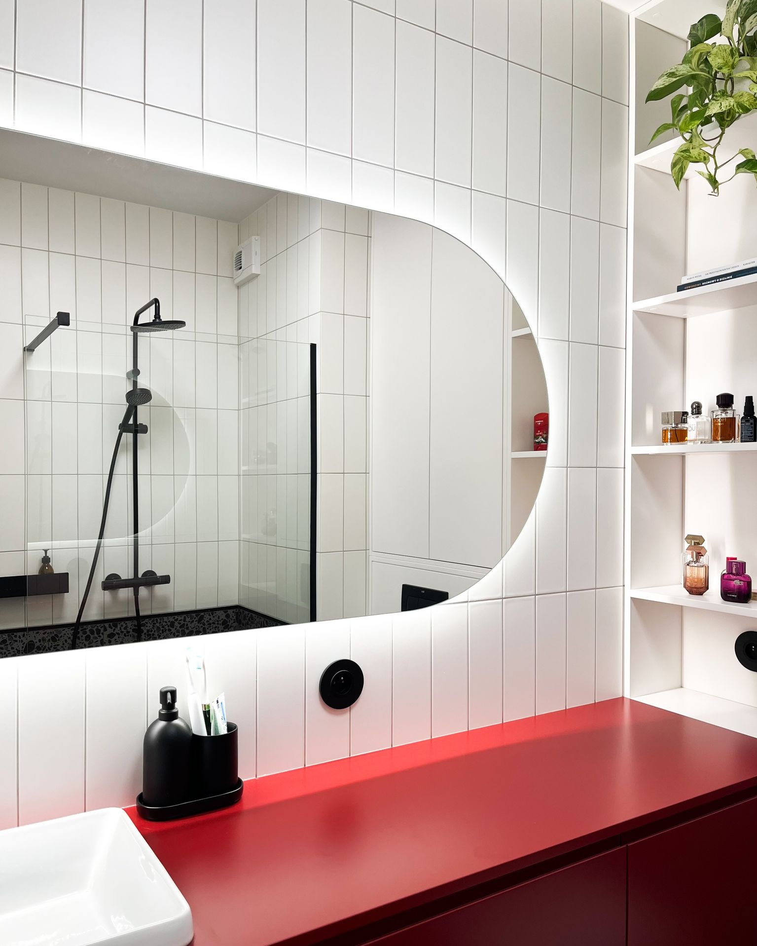 Nowoczesna łazienka z białymi, pionowymi płytkami, okrągłym podświetlanym lustrem, czerwonym blatem i czarną armaturą prysznicową widoczną w odbiciu.