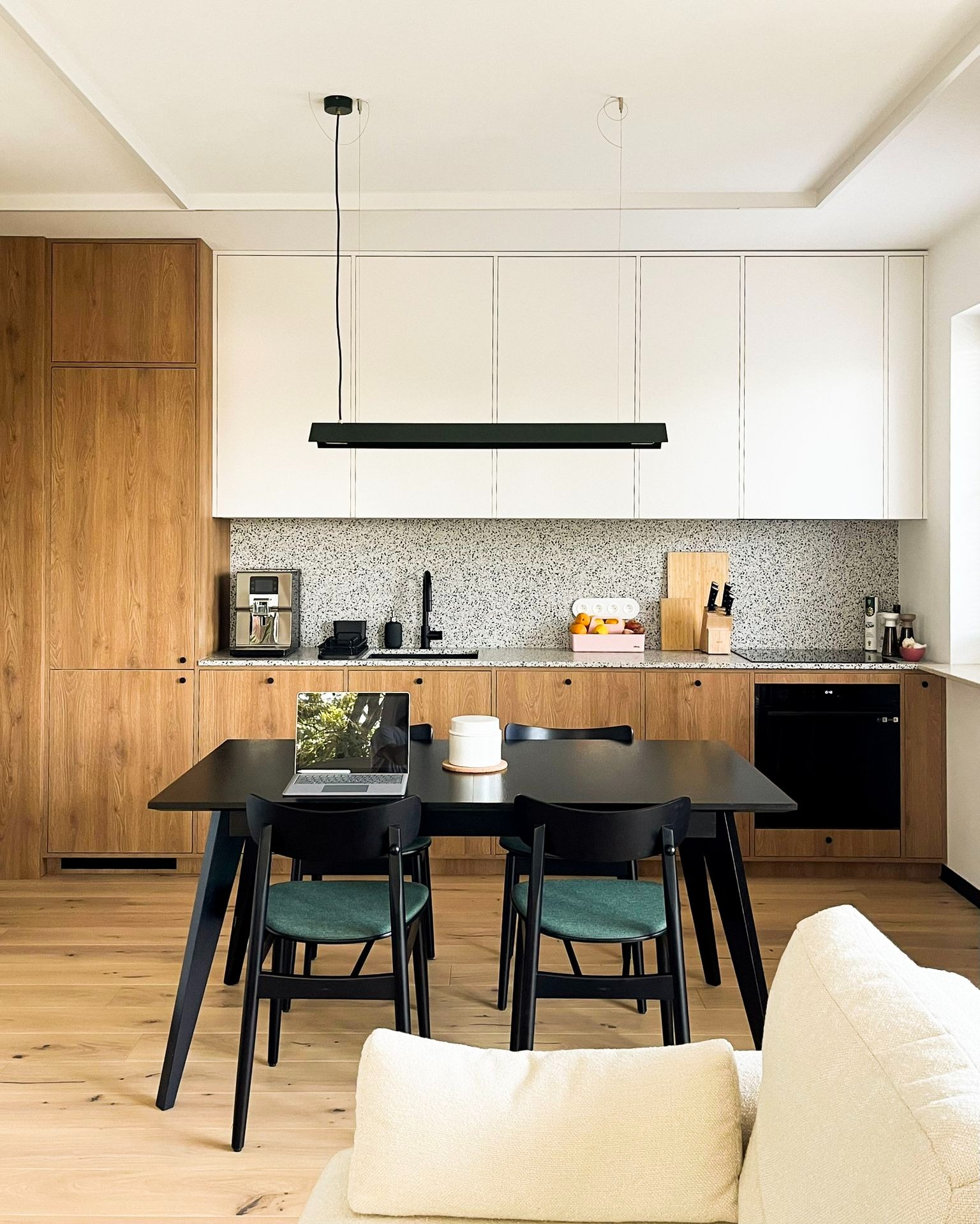 Nowoczesna kuchnia z drewnianymi i białymi szafkami, szarym blatem, czarnym stołem jadalnym i krzesłami z zielonymi siedziskami, laptop na stole, widok z salonu.