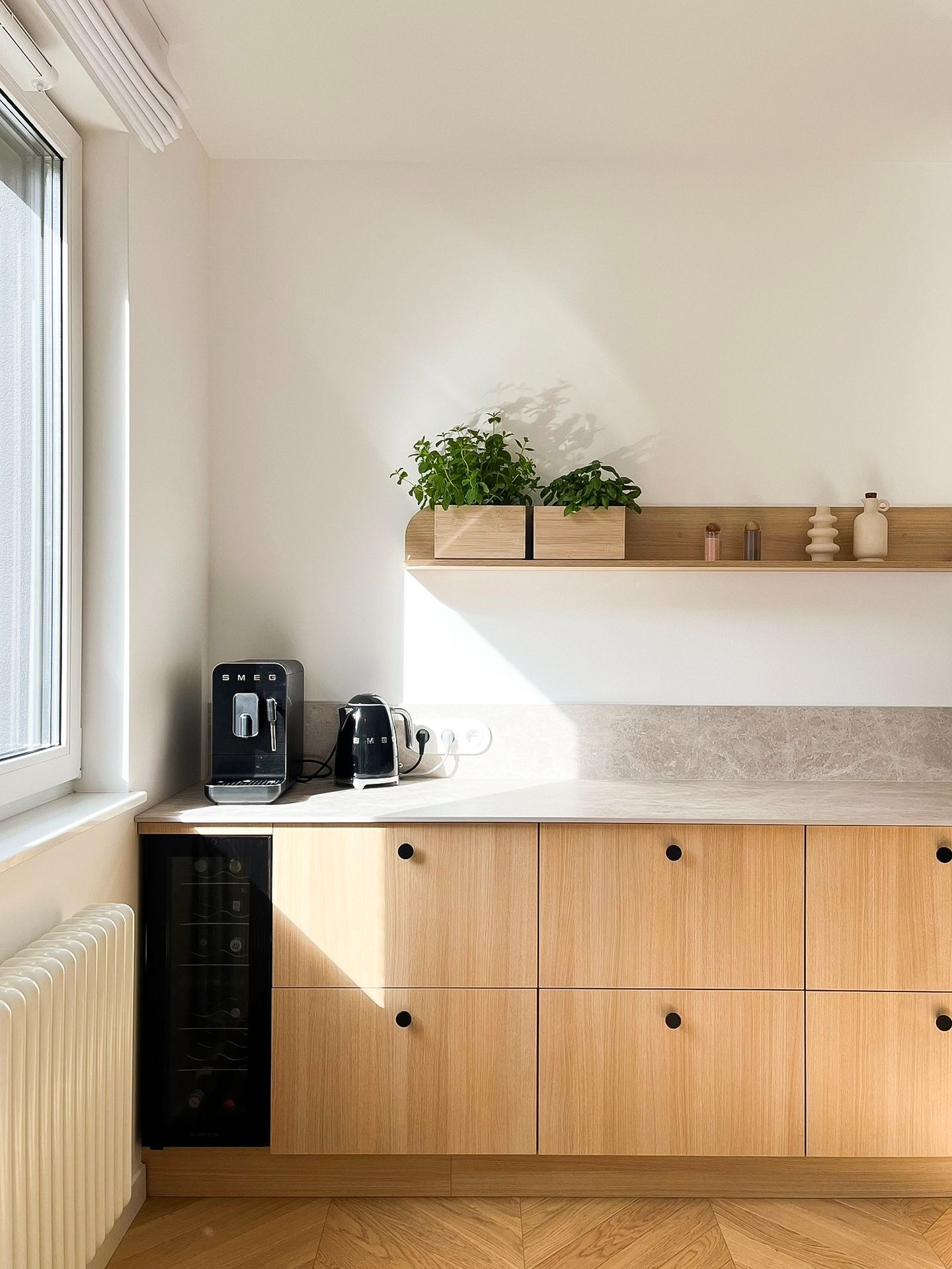 Jasna kuchnia z drewnianymi szafkami, czarnymi okrągłymi uchwytami, ekspres do kawy Smeg, czajnik Smeg, półka z ziołami i dekoracjami, parkiet w jodełkę.