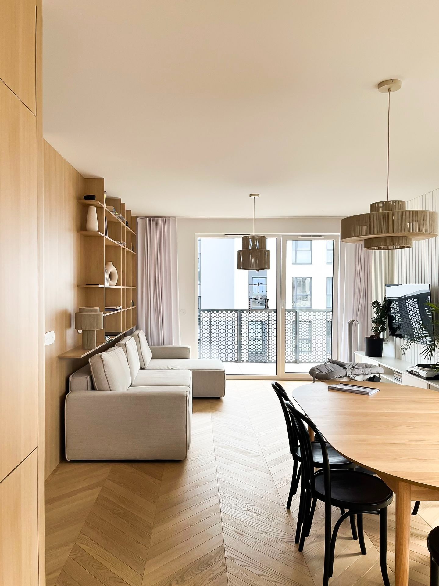 Jasne wnętrze mieszkania z drewnianą podłogą ułożoną w jodełkę, narożną sofą z półkami, okrągłym stołem z czarnymi krzesłami i lampami z abażurami ze sznurka.