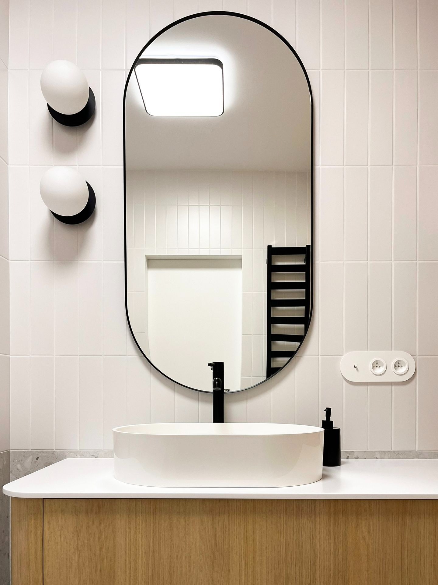 Minimalistyczna łazienka z owalnym lustrem w czarnej ramie, białą umywalką nablatową, czarną armaturą i grzejnikiem drabinkowym, oświetlona kulistymi lampami ściennymi i kwadratową lampą sufitową...