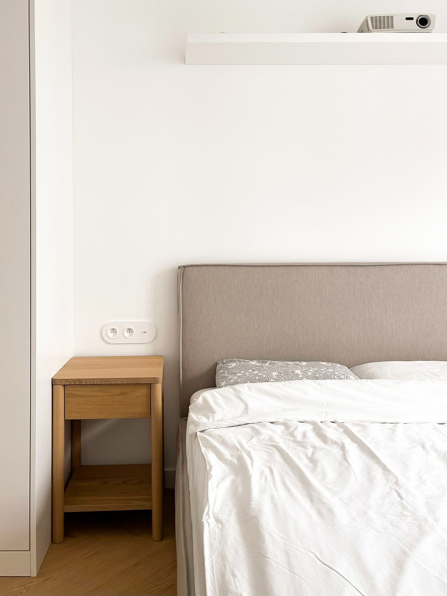 Minimalistyczna sypialnia w bieli i beżu, z drewnianą szafką nocną i projektorem na półce nad łóżkiem.