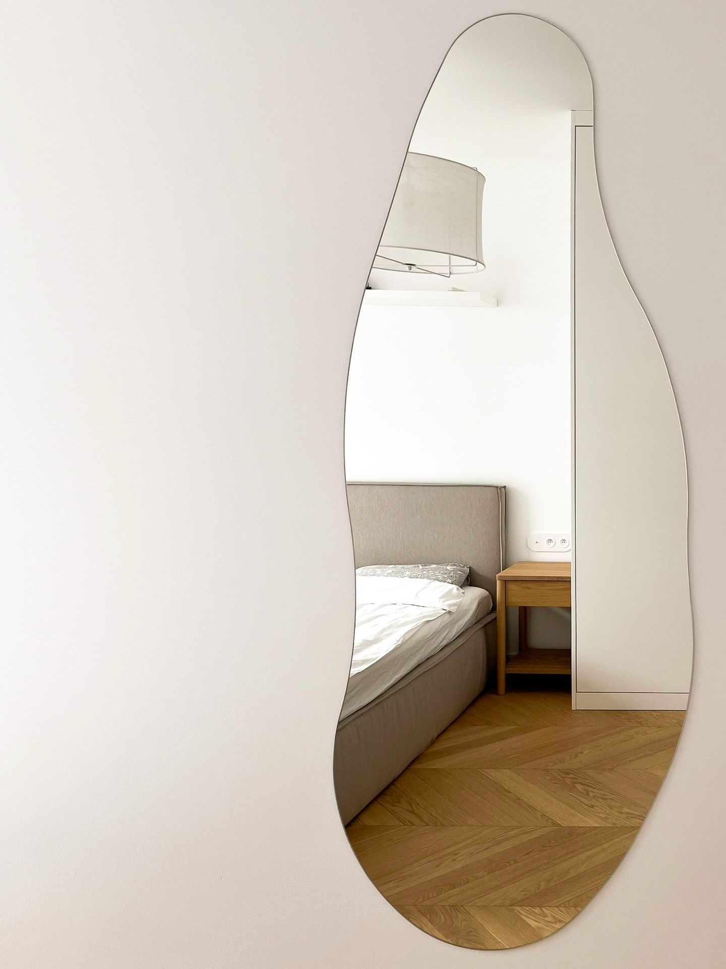 Nieregularne lustro w kształcie kropli wody odbija fragment sypialni: łóżko z szarym zagłówkiem, drewnianą szafkę nocną i parkiet w jodełkę.