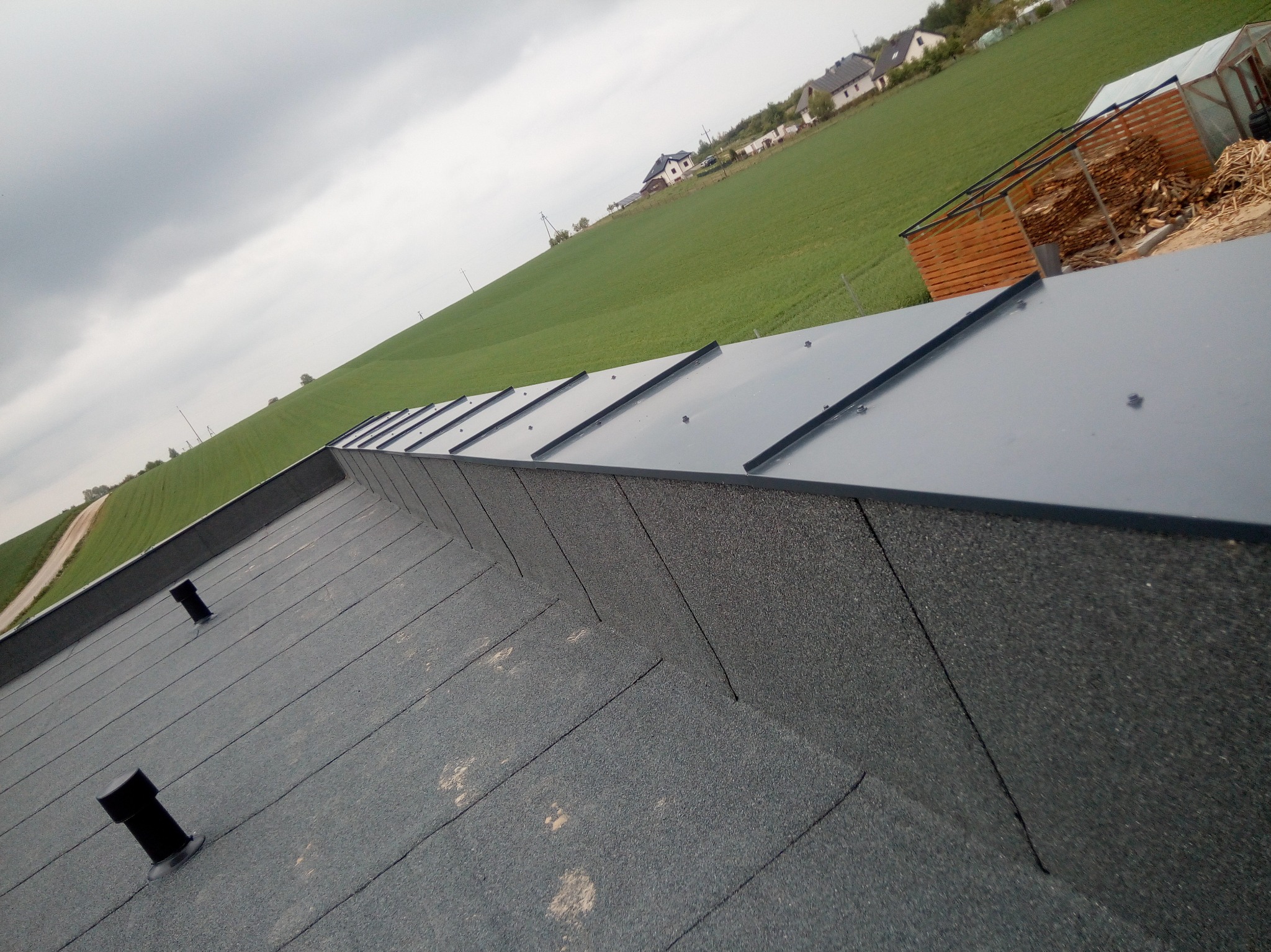 Widok z góry na płaski dach pokryty papą z kominkami wentylacyjnymi, w tle zielone pole i zabudowania.