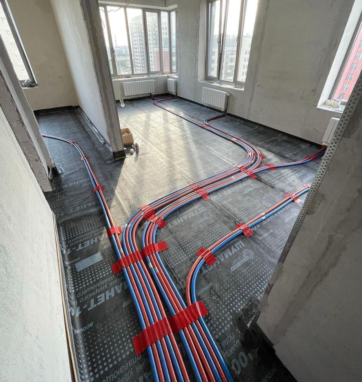 Montaż ogrzewania podłogowego: rury grzewcze, niebieskie i czerwone, ułożone na czarnej folii izolacyjnej z widocznymi napisami, przymocowane czerwoną taśmą w surowym wnętrzu z oknami i grzejnikami.
