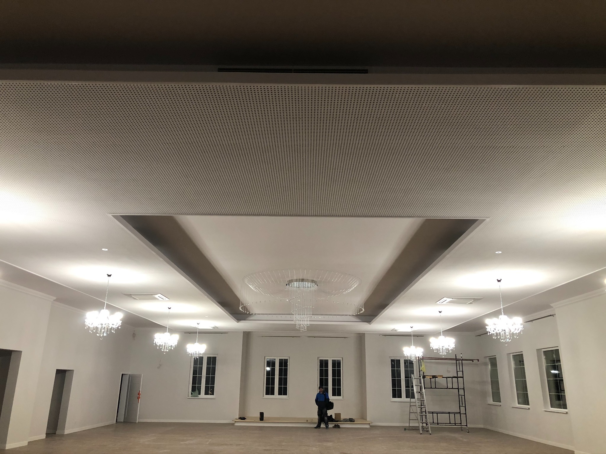 Przestronne, jasne wnętrze sali balowej z białymi ścianami, licznymi oknami, ozdobnymi żyrandolami i podwieszanym sufitem z perforowanymi panelami. Osoba w roboczym ubraniu stoi na podeście przy...
