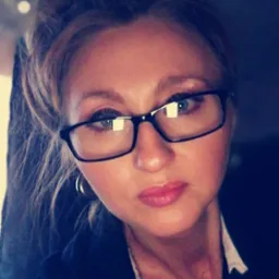 Portret kobiety w okularach, z upiętymi blond włosami i delikatnym makijażem, ubrana w ciemną marynarkę z białą koszulą