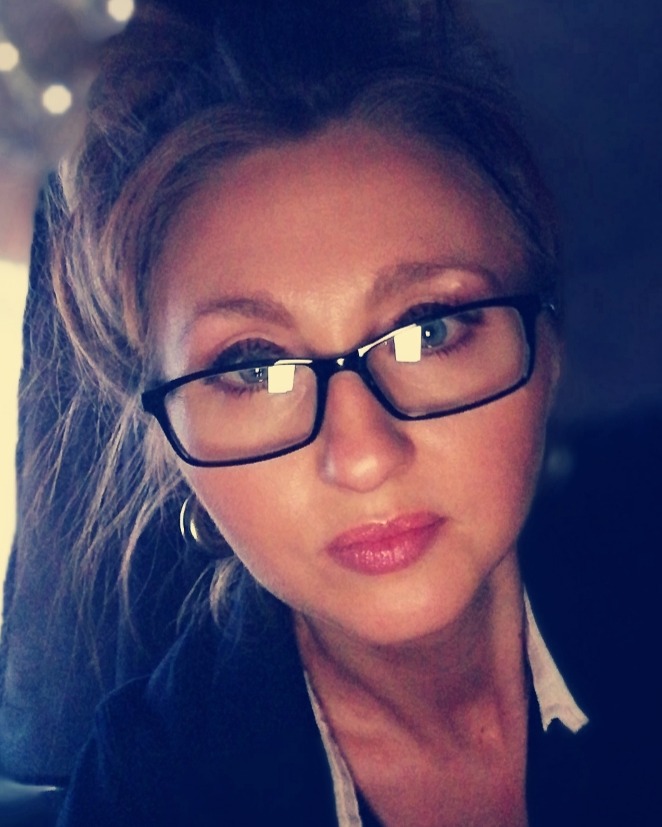 Portret kobiety w okularach, z upiętymi blond włosami i delikatnym makijażem, ubrana w ciemną marynarkę z białą koszulą