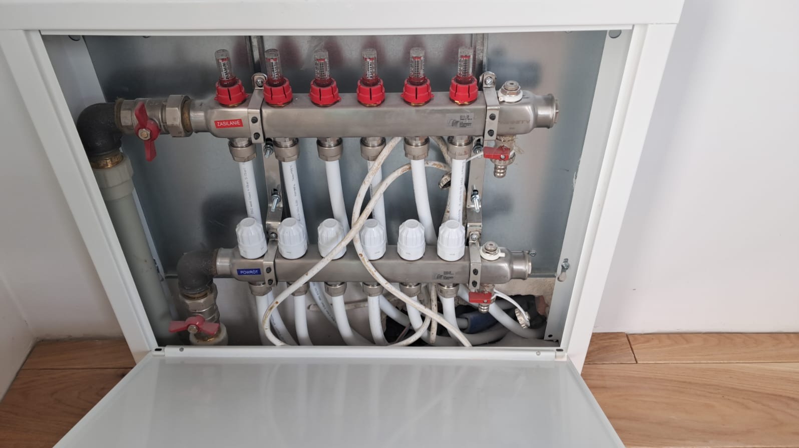 Rozdzielacz ogrzewania podłogowego w metalowej szafce, widoczne zawory z czerwonymi elementami, białe rurki i etykiety 'Zasilanie' oraz 'Powrót'.