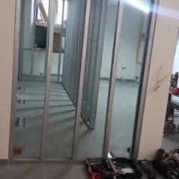 Konstrukcja z metalowych profili, tworząca szkielet ścian działowych w pomieszczeniu. Widoczne narzędzia i płyta. Etap prac przygotowawczych.