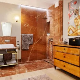 Stylowa łazienka z kabiną prysznicową wyłożoną marmurem, umywalką na złotym stelażu i komodą z obrazem. Nietypowe oświetlenie i dodatki.