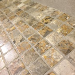 METAINVEST SPÓŁKA Z OGRANICZONĄ ODPOWIEDZIALNOŚCIĄ - Mozaika z małych, kwadratowych płytek w odcieniach beżu, szarości i brązu, ułożona na powierzchni blatu. Widoczna fuga w jasnym odcieniu. Ujęcie z bliska, pod kątem.