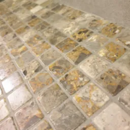 Mozaika z małych, kwadratowych płytek w odcieniach beżu, szarości i brązu, ułożona na powierzchni blatu. Widoczna fuga w jasnym odcieniu. Ujęcie z bliska, pod kątem.