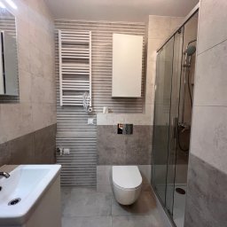 METAINVEST SPÓŁKA Z OGRANICZONĄ ODPOWIEDZIALNOŚCIĄ - Wykończona łazienka w szaro-białej kolorystyce z kabiną prysznicową, umywalką z szafką, toaletą i grzejnikiem drabinkowym. Widoczne płytki w dwóch odcieniach szarości.