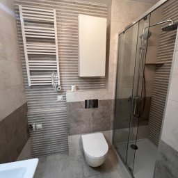 METAINVEST SPÓŁKA Z OGRANICZONĄ ODPOWIEDZIALNOŚCIĄ - Nowoczesna łazienka po remoncie: kabina prysznicowa ze szklanymi drzwiami, podwieszana toaleta, grzejnik drabinkowy i szafka wisząca na tle płytek w odcieniach szarości i beżu.