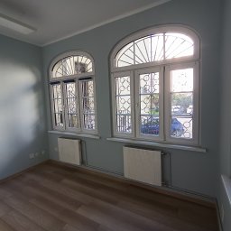 METAINVEST SPÓŁKA Z OGRANICZONĄ ODPOWIEDZIALNOŚCIĄ - Pomalowane na jasny odcień szarości wnętrze z drewnianą podłogą i dwoma oknami łukowymi z dekoracyjnymi kratami. Grzejniki pod oknami. Widok na zewnątrz.