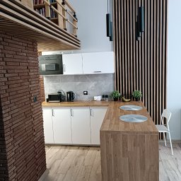 METAINVEST SPÓŁKA Z OGRANICZONĄ ODPOWIEDZIALNOŚCIĄ - Nowoczesna kuchnia z ceglanym filarem, drewnianym blatem i lamelami na ścianie. Widoczne białe szafki, antresola z książkami i designerskie lampy.