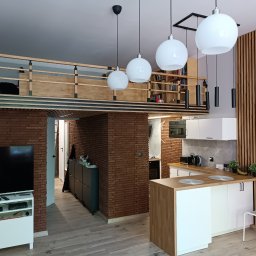 METAINVEST SPÓŁKA Z OGRANICZONĄ ODPOWIEDZIALNOŚCIĄ - Nowoczesne wnętrze z antresolą, ceglaną ścianą i drewnianymi lamelami. Biała kuchnia z wyspą i okrągłymi lampami. Widoczny telewizor i regał z książkami.