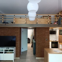 METAINVEST SPÓŁKA Z OGRANICZONĄ ODPOWIEDZIALNOŚCIĄ - Nowoczesne wnętrze z antresolą i ceglaną ścianą. Widoczny salon z telewizorem, kuchnia z drewnianym blatem i oświetlenie. Całość utrzymana w jasnej kolorystyce.