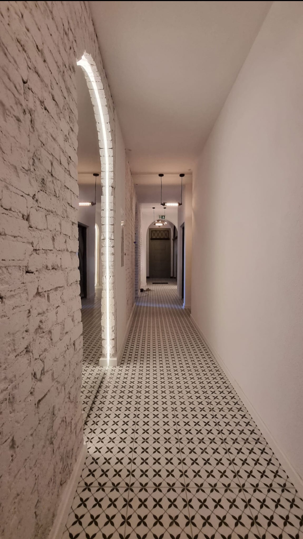 Długi korytarz z białą cegłą i podświetlonym łukiem, wzorzysta podłoga, minimalistyczne lampy. Perspektywa z głębi korytarza, jasne ściany.